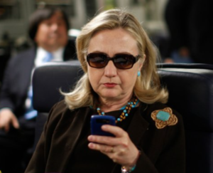 Hillary Clinton seated on a plane, looking at her cellphone, wearing sunglasses.
